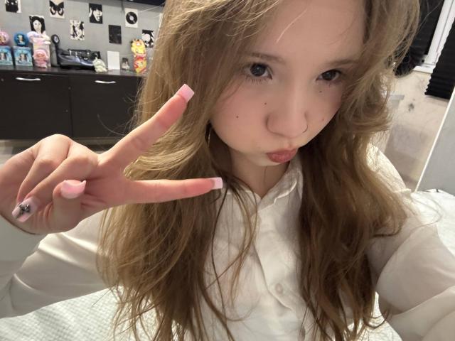 Close-up photo of YanDelangel with long brown hair, making a peace sign with pink-manicured fingers, pouting lips, wearing a white shirt, in a brightly lit room with black cabinets and butterfly decorations.