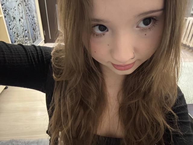 Photo en gros plan de YanDelangel, avec de longs cheveux bruns, des yeux bleus et un anneau de nez, regardant directement l'appareil photo. Expression légèrement boudeuse. Elle porte un haut noir. Incroyablement sexy..