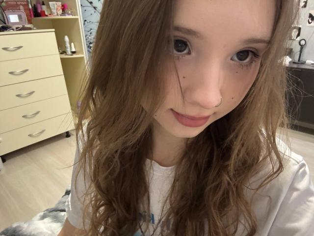 Close-up of YanDelangel with long, wavy brown hair, pale skin, and blue eyes, looking directly at the camera with a subtle smile. Background shows a cluttered room with a dresser. Fucking cute.