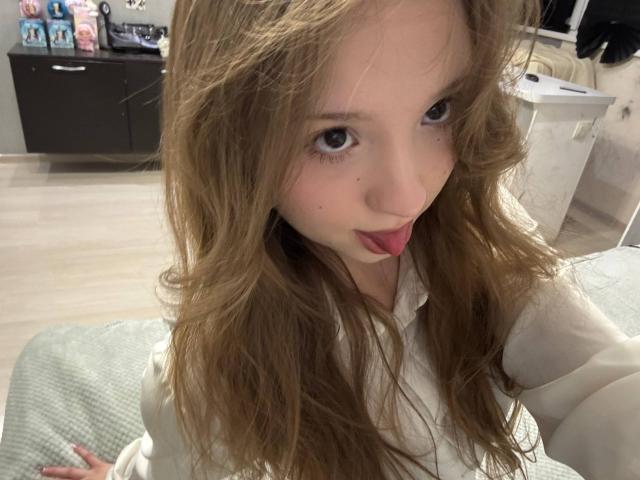 Primer plano de YanDelangel con cabello castaño largo y ondulado, sacando la lengua de forma provocativa. Ella tiene piel clara, ojos azules y lleva una camisa blanca. El fondo incluye estanterías y objetos borrosos. Tensión sexual.