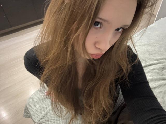 Close-up selfie van YanDelangel met lang, rommelig bruin haar, een intense blik en lichtjes open lippen. Ze draagt een zwarte top, leunt voorover, en er is een deel van haar geslachtsdeel zichtbaar tussen haar benen..