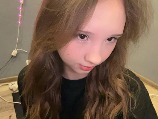 Close-up of YanDelangel, a young woman with fair skin and long, wavy brown hair, wearing a black top. She has a seductive gaze and slightly parted lips. Dimly lit room.