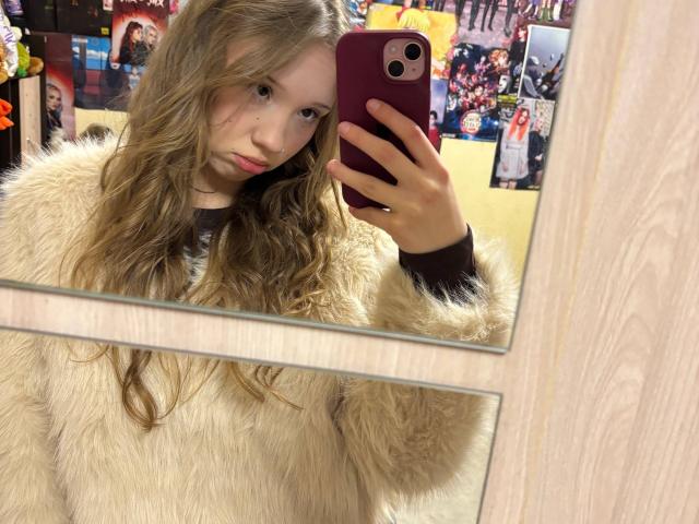 YanDelangel, avec de longs cheveux bruns ondulés, portant un manteau beige duveteux, tenant un téléphone violet, se prend en photo dans un miroir. Contexte : magasin coloré et encombré, avec des affiches et des présentoirs. Tension sexuelle : Le regard subtil et séduisant de YanDelangel..