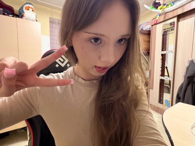 YanDelangel, young woman with long brown hair, pale skin, and small breasts, making a peace sign with pink nail polish, slightly open mouth, beige shirt, bedroom setting, cluttered closet.