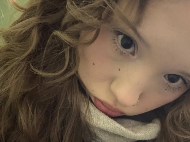 Close-up of YanDelangel with wavy brown hair, blue eyes, and several facial moles. YanDelangel's face is tilted slightly to the side, with a relaxed, almost seductive expression. Gray background.