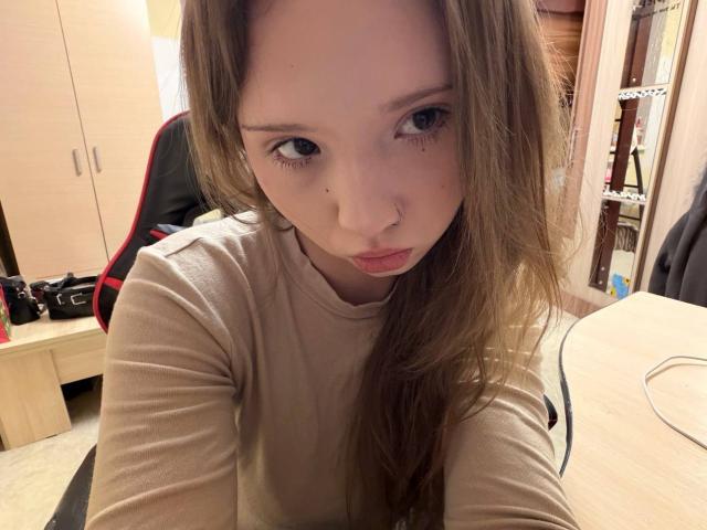 Primer plano de YanDelangel con cabello largo y castaño, mirando directamente a la cámara con una expresión seria. Ella lleva una camisa color beige y tiene el cartílago de la nariz perforado. El fondo muestra un escritorio y una silla desordenados..