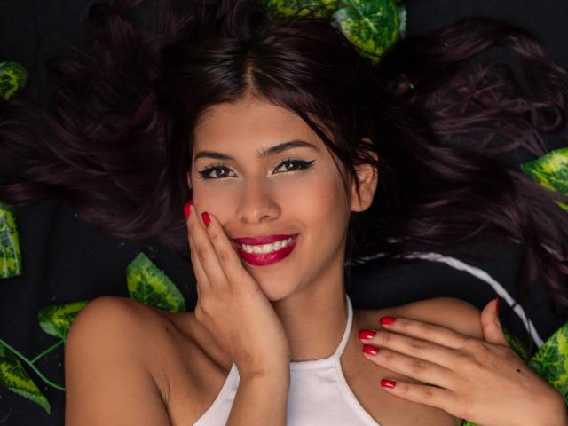 Foto em close-up de DanikaSerov, deitada de costas com o cabelo escuro espalhado, vestindo uma blusa branca de alças e com unhas vermelhas. Sorrindo, com batom vermelho, e folhas verdes ao redor da cabeça. Muito atraente..