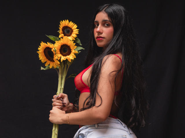 TiffannyRose, con lunghi capelli neri, indossa un reggiseno rosso e pantaloni bianchi, e tiene in mano un bouquet di girasoli. Ha un'espressione sensuale, uno sfondo nero, colori vivaci di rosso e giallo..