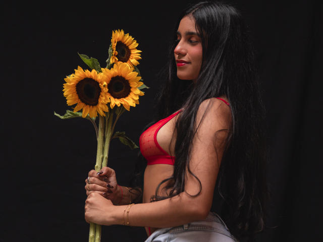 TiffannyRose, a curvy woman with long black hair, holds a bouquet of sunflowers against a black background. She wears a red bra, white pants, and has a seductive smile.