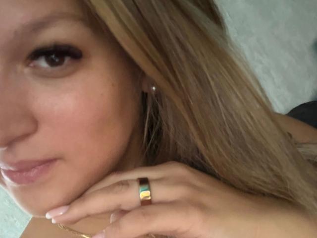 Close-up van het gezicht van Elli69-hot, met blond haar, lichte huid en subtiele make-up. Ze kijkt in de camera, met haar hand in de buurt van haar kin, en draagt een groene ring aan haar vinger..