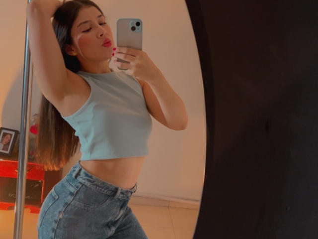 EmmaLombardi takes a mirror selfie, arm raised, wearing a tight blue crop top and high-waisted jeans. She's sucking on her finger, eyes closed, with a sexy, confident pose. Smartphone in hand.