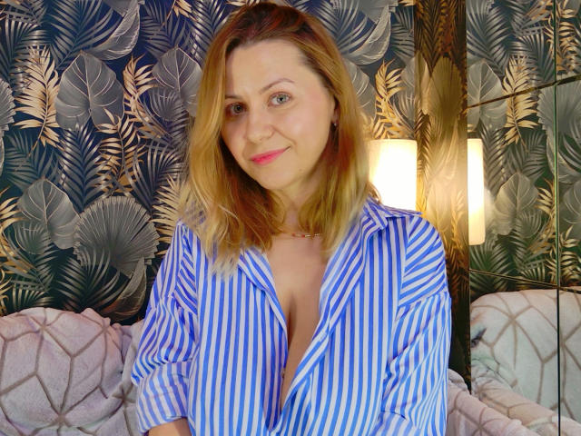 NymphoArri, blonde, medium-length hair, blue eyes, wearing an unbuttoned blue and white striped shirt, slight smile, standing in front of a leaf-patterned wall, soft light, subtle sexual tension.