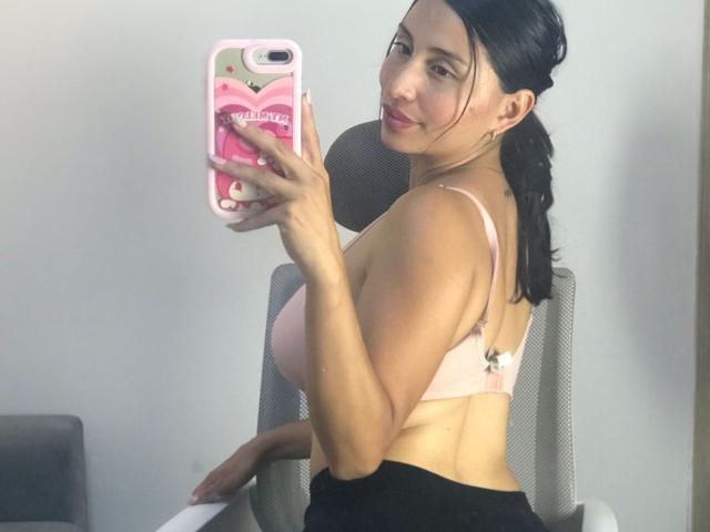 Kate Owens si fa un selfie allo specchio, tenendo in mano un telefono rosa con una custodia rossa. È senza top, mostrando il suo seno prosperoso, con i capelli scuri raccolti in una coda di cavallo e pantaloni neri..