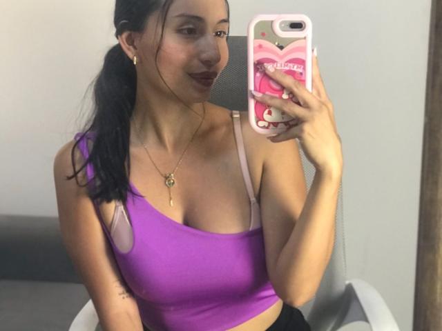 KateOwens, brunette with long hair in ponytail, wearing a tight purple crop top with pink straps, holding phone with pink case, taking mirror selfie, subtle smile, gold necklace, visible cleavage.