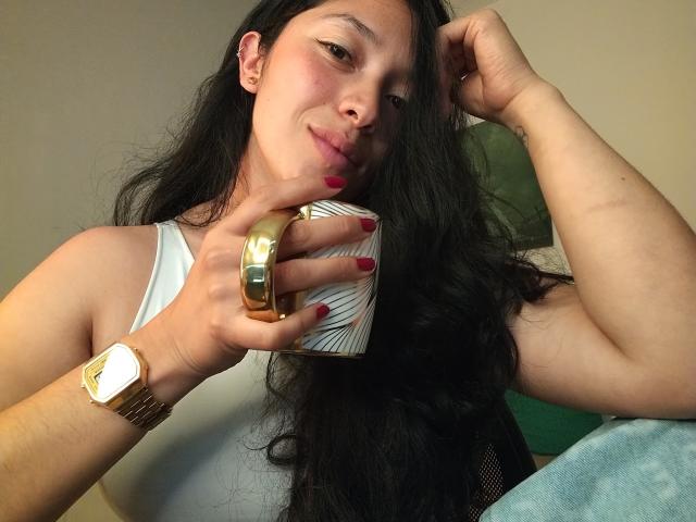 ChantalBernadette, uma mulher asiática atraente com longos cabelos pretos, segurando um anel de ouro na mão direita e uma xícara branca em sua mão. esquerda. Ela veste uma blusa branca e um relógio dourado. Incrivelmente sexy..