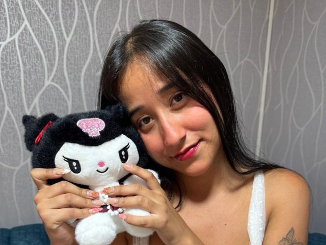 AnnieBlakke tient un chat en peluche noir et blanc, avec un nœud rose, ses cheveux noirs sont lâchés, et elle sourit légèrement. Elle porte un débardeur blanc, et l'arrière-plan présente un papier peint à motifs ondulés..