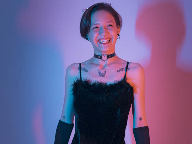 LissyLuxe è al centro, sorridente, indossa un abito nero con piume, un collare e guanti neri. L'illuminazione blu e rosa al neon proietta ombre drammatiche. I suoi capelli castani corti sono leggermente spettinati. Trasmette una forte carica erotica..