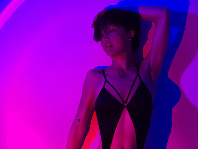 LissyLuxe, kort donker haar, zwarte bandeau top, arm omhoog, verleidelijke pose, levendige neon roze en blauwe verlichting, schaduwen op het lichaam, tatoeage op de rechterarm, sexy, zelfverzekerde uitdrukking, sfeer van een nachtclub..