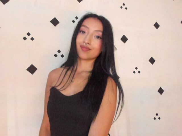 Photograph of HotMoonX with long black hair, wearing a black tank top, standing against a white wall with black diamond patterns. She has a confident smirk, sexy look, and slight forward lean.