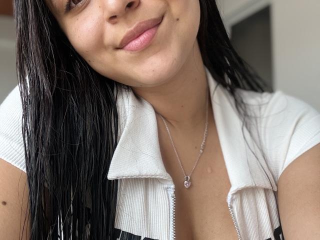 Close-up photo of HikariChloe, light-skinned woman with long black hair, slightly tilted head, pink lips, small mole on cheek, white collared shirt, silver necklace with pendant, subtle smile, sexual tension.