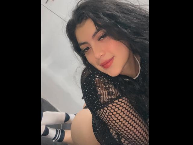 Close-up de MiaaAmour, uma mulher atraente com cabelos longos e pretos, vestindo uma blusa de rede preta, olhando sedutoramente para... câmera. Ela tem pele clara, lábios cheios e uma expressão confiante..