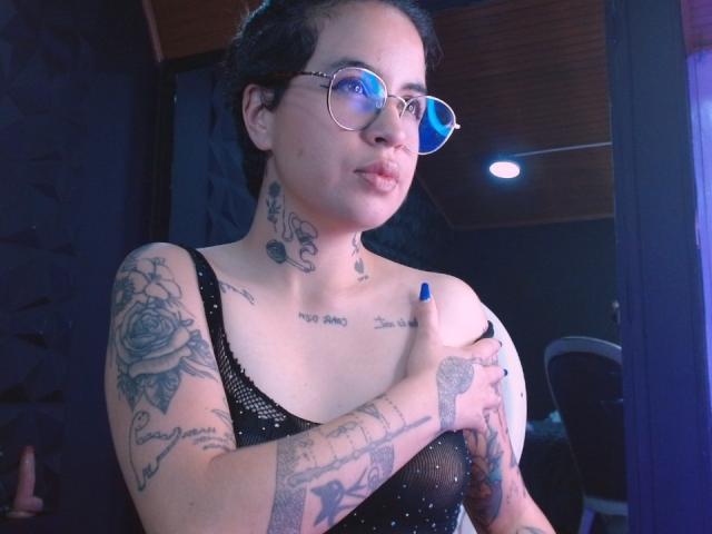 Photograph of SamyEvanss, tattooed woman with short black hair, wearing glasses and a black mesh top, holding her small breasts with a hand, blue-tinted lighting, sexy and confident pose.