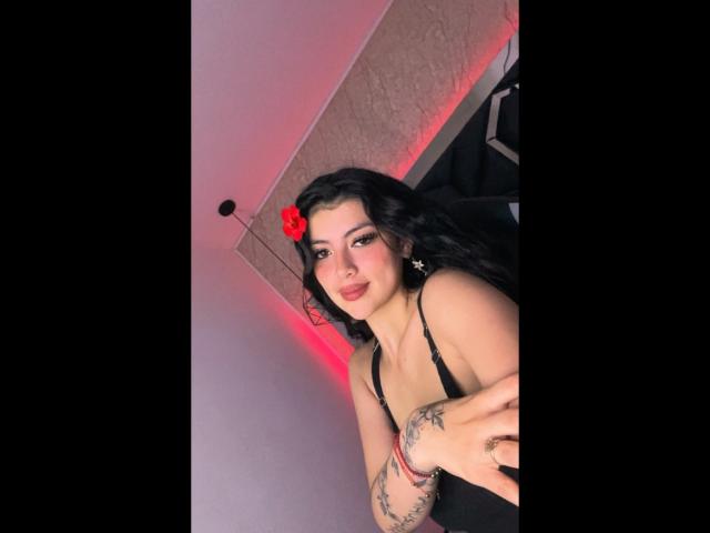 Digitaal portret van MiaaAmour, een sexy bruinharige vrouw met zwart haar, een rode bloemclip, een zwarte jurk met bandjes en tatoeages op de onderarmen. Voorovergebogen, verleidelijke blik, rode en roze verlichting, intense seksuele spanning..