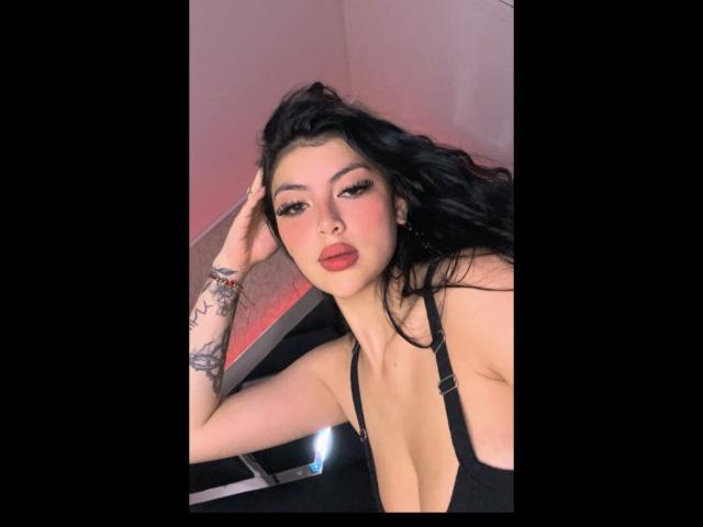 Fotografia di MiaaAmour con pelle chiara, capelli neri e rossetto rosso, sdraiata, con una mano tra i capelli, e scollatura nera che mette in risalto il seno. donna, tatuaggi sul braccio sinistro, sguardo intenso, illuminazione rosa tenue.