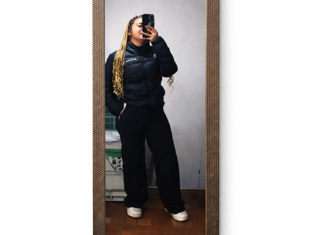 Tatiana Rossi, una mujer de talla grande con largas trenzas rubias, está de pie frente a un espejo, tomando una selfie con su teléfono. Ella lleva una chaqueta negra acolchada y pantalones negros, zapatillas blancas..
