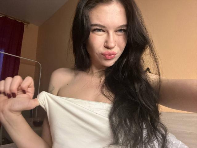 NgaKrahn, con cabello negro largo, labios rosados y apretados, tirando de una correa blanca, mostrando sus pequeños pechos, en un dormitorio, con un tatuaje en el hombro izquierdo..