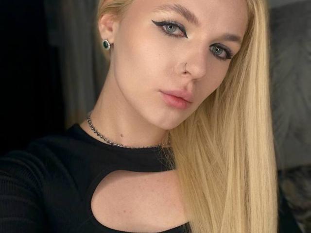 Gros plan de MichelleHoock, une femme blonde aux yeux bleus, portant un eye-liner noir et un haut noir avec une découpe en forme de clé. Elle a un anneau au nez, des boucles d'oreilles et un sourire subtil. Putain, elle est sexy..