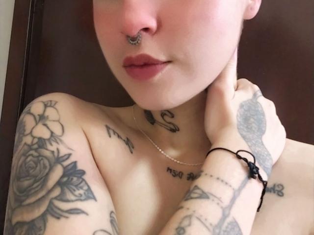 Close-up photo of SamyEvanss, light-skinned with tattoos, nose ring, and black rose tattoo on shoulder. SamyEvanss' hand touches their neck, showing black bracelet. Visible collarbone tattoos. Sexy, suggestive pose.