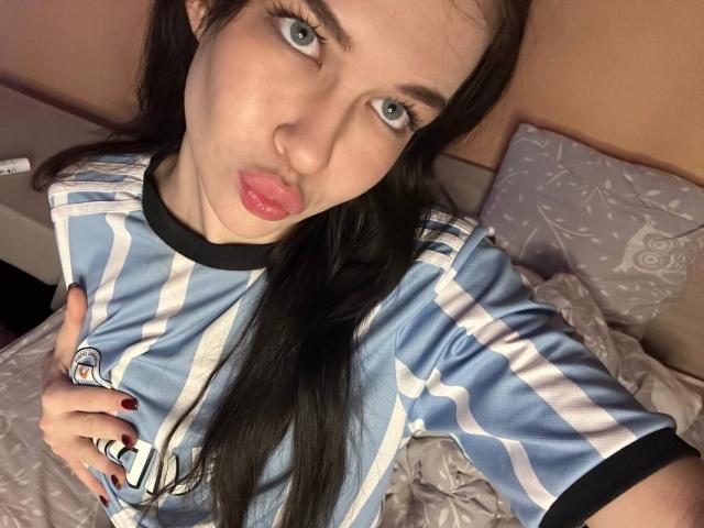 NgaKrahn, con labios rosados y fruncidos, camiseta de fútbol a rayas azules y blancas, cabello negro largo y uñas negras, sosteniendo el pecho, ojos verdes, mirando a la cámara..