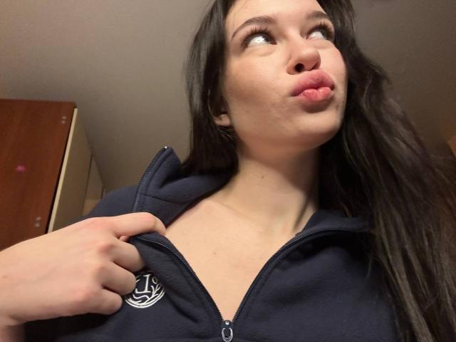 NgaKrahn, avec ses lèvres boudeuses, ses longs cheveux noirs et sa peau claire, zippant une veste à capuche bleu marine, une main sur la fermeture éclair, regardant vers le haut avec une expression espiègle..
