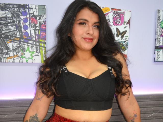 Greace Wattson, avec ses longs cheveux noirs ondulés, portant un soutien-gorge noir, est assise devant une œuvre d'art abstraite. Elle a des tatouages sur les bras, et un sourire léger et confiant. Incroyablement sexy.