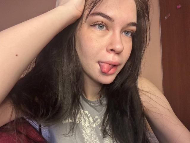 NgaKrahn with long brown hair, blue eyes, and fair skin, sticking out her tongue, right hand in hair, wearing a grey shirt, in a casual selfie.