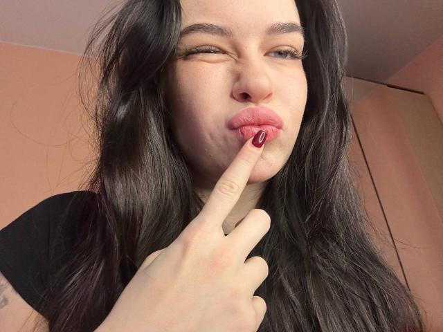 NgaKrahn, avec ses longs cheveux noirs, cligne de l'œil, fait la moue avec son rouge à lèvres rose, et porte un doigt sur sa lèvre inférieure. Elle a la peau claire, porte une chemise noire et a les ongles rouges..