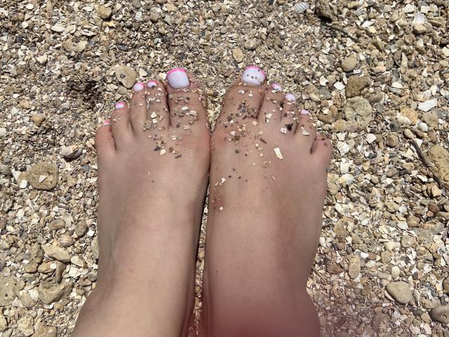 Los pies de Barbarica, con las uñas de los pies pintadas de rosa, están sobre una playa de guijarros. Granos de arena se adhieren a la piel. Los pies, carnosos y de color rosado-marrón, contrastan con las piedras blancas. Primer plano explícito, tensión sexual, ambiente de playa..