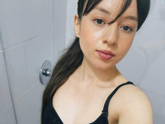 Close-up van VioletaNova, een jonge Aziatische vrouw met natte huid, zwart haar in een paardenstaart en roze lippen, die een zwarte tanktop. Haar blik is direct en verraadt een subtiele seksuele spanning..