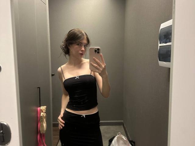 NgaKrahn mit kleinen Brüsten in einem schwarzen, trägerlosen Top und einem passenden schwarzen Rock, macht ein Selfie vor einem Spiegel in einem schwach beleuchteten Raum und hält dabei ein iPhone mit einer rosa Hülle..