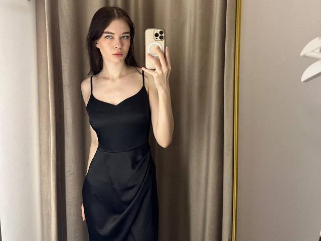 NgaKrahn, con cabello largo y castaño, vestida con un vestido negro, sosteniendo un iPhone, tomando una selfie frente al espejo, mostrando sus pechos y glúteos, de pie frente a cortinas beige..