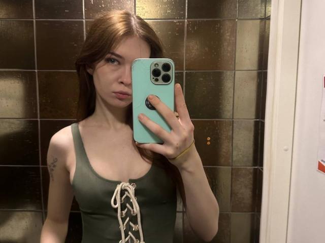 NgaKrahn with red hair, green tank top, lace-up front, holding a blue phone, mirror selfie, visible tattoo on left arm, yellow bracelet, serious expression, brown tiled wall.