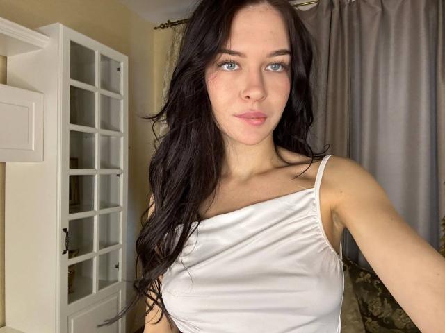 NgaKrahn with long black hair, blue eyes, and a white tank top showing small tits, standing in a room with a white door and gray curtains.