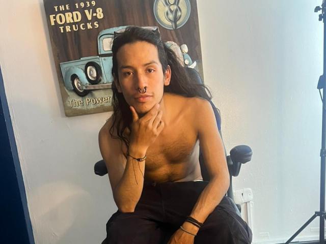 EnzoJagger, senza camicia, seduto su una sedia, con lunghi capelli scuri, pelle marrone, corporatura esile, pantaloni neri, una mano sul mento, macchina fotografica. un poster sullo sfondo, luce soffusa, tensione sessuale, posa rilassata, braccialetti..