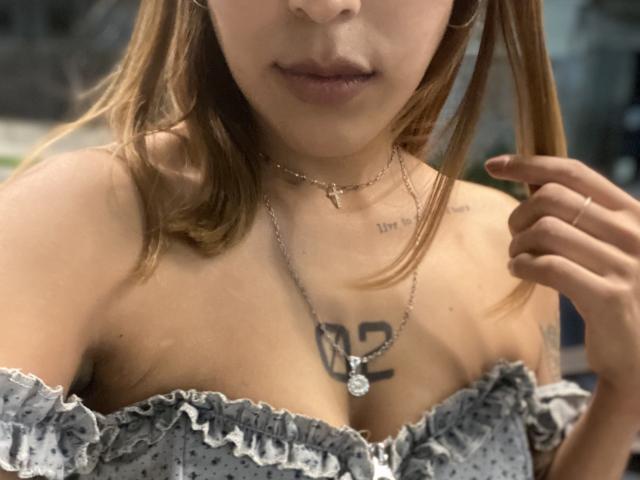 Nahaufnahme von KiaraHernandezX, wobei der Fokus auf ihrem Dekolleté liegt. Sie trägt ein Top mit einem Schulterfrei-Schnitt und Rüschen, und man kann Tattoos auf ihrem Körper sehen. Brust, mit einer Halskette, hält ihr Haar, zeigt leichte sexuelle Anspannung..