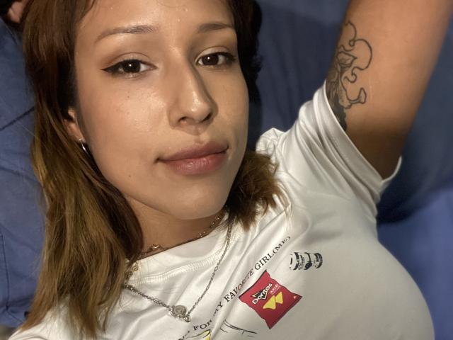 Close-up of KiaraHernandezX with light brown skin, brown eyes, and shoulder-length brown hair. She's wearing a white T-shirt, has a tattoo on her right arm, and lying on a blue surface. Her expression is relaxed and slightly seductive.