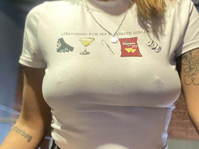 Nahaufnahme von KiaraHernandezX, die ein weißes T-Shirt mit dem Aufdruck "Alles für meinen Lieblingsapfelwein" trägt, wobei ihre großen, sichtbare Brustwarzen durch den Stoff. Sie hat Tattoos an ihren Armen. Sexuelle Spannung.