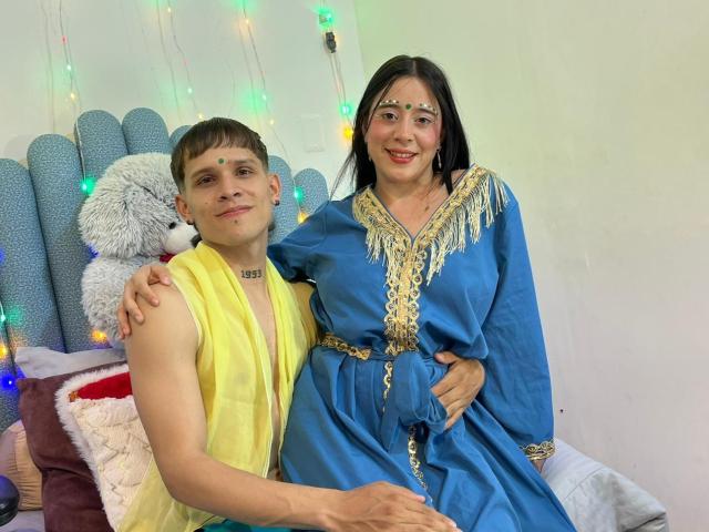 AnjaliRohan, con i capelli castani corti e vestita di giallo, seduta accanto a una donna sorridente con i capelli neri e vestita di blu. con bordi dorati. Lui le sta stringendo la vita, lei gli sta toccando la spalla. Luci colorate a forma di filo sullo sfondo..