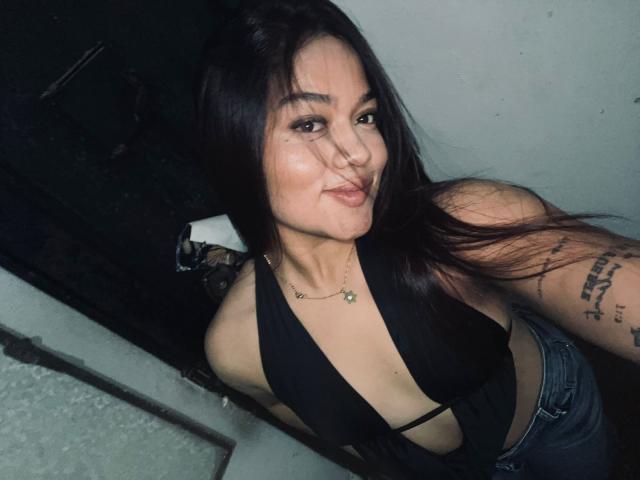 ChanneleCollins, com longos cabelos pretos, vestindo uma blusa preta de alças, mostrando uma tatuagem no braço direito, apoiada em um superfície escura, olhando diretamente para a câmera com um sorriso discreto.