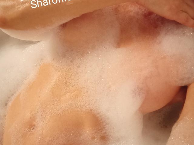 Close-up of SharonLeidy's wet, orange-toned body covered in white foam. Focus on her large, round breasts and crotch, with visible skin texture and slight glistening. Steamy, sensual atmosphere.